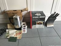 The lot on floor showing the Black & Decker steamer box, stainless steel coffee percolator with black handle and lid, set of stainless steel knives in stand, and instruction/cleaning papers.