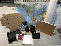 View of lot items arranged together including a laminated world map, cardboard boxes, cork board, desk lamps, file organizer, paper rolls, pad of sticky notes and loose leaf paper.