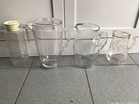 Set of 4 glass pitchers of different sizes and shapes arranged on tiled floor against white wall