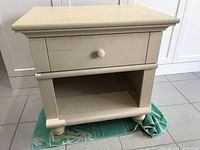 Full view of beige wooden side table showing drawer with knob and open lower shelf on four rounded feet, photographed on tiled floor with green mat underneath.