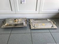 Two stainless steel serving trays side-by-side on floor, one with lid, both with copper floral inlay and handles.
