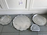 Overview of four trays including two large handled aluminum trays, one smaller round tray, and a small scalloped tray, all embossed with pine cone patterns.