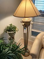 Full view of the floor lamp showing beige shade, metal base, and stand amid room plants and furnishings.