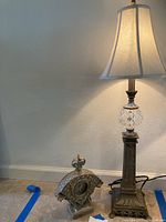 Full view of brass and glass table lamp illuminated with white fabric lampshade, beside ornate brass table clock on floor.