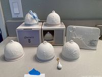 Photo showing five Lladro porcelain bells, two with boxes beneath them, and a signed decor plaque to the right. A small tag or blue tape note is partially visible.