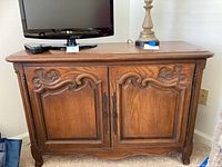 Front view of wood cabinet showing carved decorative panels on double doors and top surface holding separate lamp and TV items.