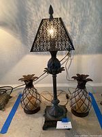 Full front view of the lamp lit with two pineapple candle holders on either side, showing the decorative shadows from the lamp shade and details of the items.