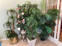 Front view of several silk trees and artificial plants grouped together near a wall and a window, showing detailed foliage and floral arrangements.
