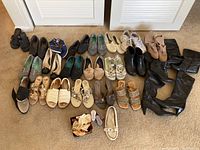 Photo showing a variety of women's shoes and boots arranged loosely on carpet including knee-high boots, ankle boots, sandals, sneakers, and dress shoes in multiple colors and styles.