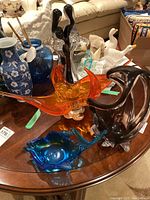 Four glass items displayed on a wooden table including blue fish-shaped bowls and a black and orange glass couple figurine.