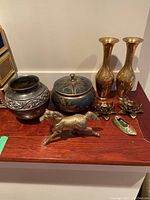 Overview of all metal decorative items including brass vases, candle holders, horse figurine, pewter vase and lidded bowl on wooden surface.