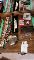 Overview of beaded necklaces, pins, watch, sunglasses, silver heart-shaped jewelry box, and bracelet on a wooden surface.