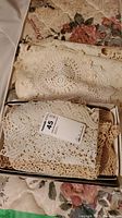 Box containing several folded crocheted doilies showing intricate lace patterns in cream color placed on floral fabric