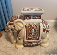 Left side angled view of decorative ceramic elephant plant stand showing detailed brown and blue ornamental designs and flat square top surface with some sticker residue.