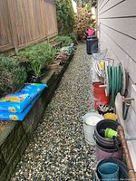 View down pathway showing bags of unopened potting soil, multiple plastic and ceramic pots, hose reel with hose, and some green plants.
