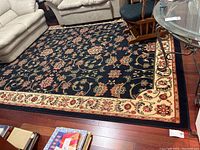 Full view of the navy floral traditional area rug with cream border on a wood floor surrounded by furniture.