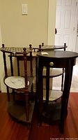 Photo showing two dark wood corner shelves with white crocheted doilies and two side tables, one round with drawer, one small with drawer, all with visible scratches.