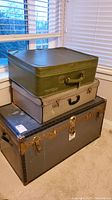 Photo showing three stacked vintage luggage pieces: a green suitcase on top, a textured suitcase in the middle, and a large steamer trunk at the bottom.
