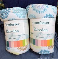 Two rolled, packaged comforters standing upright showing size labels and pattern color samples.