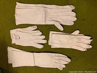 Top-down view of four pairs of white ladies' kid leather gloves on green surface. Different lengths and styles visible.
