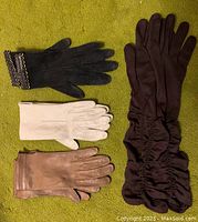 Four pairs of vintage ladies gloves laid flat on green fabric, showing black, white, tan kid leather gloves and mid-length brown nylon gloves.
