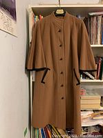 Front view of a ladies tan wool coat sized 10 in excellent condition, showing black trimmed pockets, black buttons, and shawl style long cape sleeves.