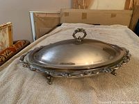 Large ornate silver plated covered casserole dish closed and resting on cloth surface.
