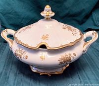 Full view of the covered white porcelain tureen with gold floral decoration and trim, showing handles and finial knob on lid.