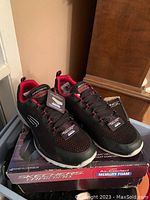 Pair of new Skechers training shoes, black with red trim, with tags on top of Skechers branded shoe box.