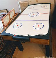 Full view of the hockey table showing white playing surface with red and blue rink markings, wood-finish frame, and black legs, positioned in room with surrounding furniture.
