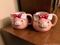 Two pink pig-shaped mugs with red polka dot bows showing front face and handles