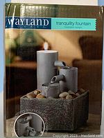 Front side of box showing the fountain with water flowing from cylindrical spouts into a bottom square base filled with faux rocks and a lit tealight on top.