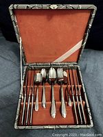 Flatware set arranged inside open vintage box with red velvet lining, showing knives, spoons, forks, and accessory pieces.