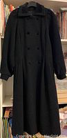 Front view of the woman's long black wool coat, showing double-breasted button closure, pockets, bell sleeves, and full length.