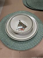 Close-up showing stacked plate, bowl with Christmas tree design, and green-gold woven placemat underneath.