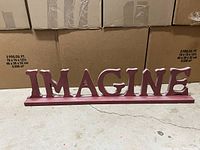 Front view of the decorative word art spelling 'IMAGINE', showing distressed white letters on a dark red base.