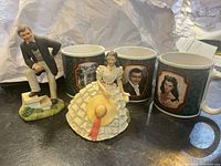 Front view of Rhett Butler and Scarlett O'Hara figurines with three collectible mugs behind them showing character portraits from Gone With The Wind.