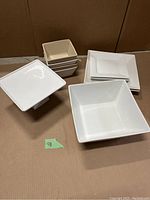 Photo showing all square white ceramic pieces including bowls, plates, and cake stand arranged on cardboard.
