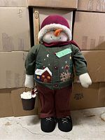 Front view of plush snowman showing facial features, winter hat, scarf, green jacket with appliques, maroon pants, black feet, and bucket with plush snowballs in hand.