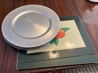 Stack of round plastic chargers on top of six rectangular placemats with fruit artwork and green borders on a wooden table.