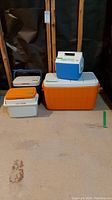 Photo showing four coolers grouped: two large Coleman coolers orange/white, one small blue and white Igloo cooler, one small orange and white cooler