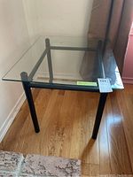 Side angle of the glass top table on wooden floor showing metal legs and glass top.