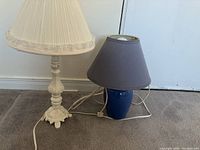 Two table lamps shown side by side. One lamp has ornate cream carved base and cream pleated lampshade, the other has blue ceramic base and gray shade. The lamps have cords and plugs visible.