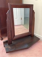 Front view of rectangular wooden framed mirror with tilting supports and wooden base shelf on carpet.