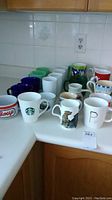 Photo showing assortment of mugs on kitchen counter, including white ceramic mugs, blue mugs, patriotic bowl-shaped mug, Starbucks mug, and mugs with animal and letter designs.