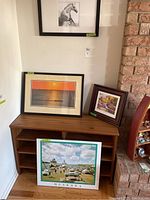 Four framed pictures displayed on and above a wooden console: a black and white horse print, muskoka beach scene print, sunset over water, and small street scene framed picture