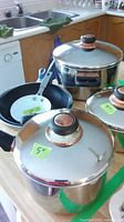 Photo showing three stainless steel pots with copper color handles on lids and a black frying pan with a white sticker on handle.