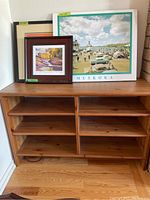 Front view of wooden shelving unit with adjustable shelves and scratches. Several framed artworks placed on top.