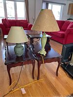 Two table lamps side by side on wooden tables with visible power cords and fabric lampshades, one lamp has a carved light green base, the other has a round dark green base.