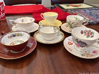 Set of six assorted vintage teacups with saucers arranged on table, showing various floral patterns, gold trims, and different cup shapes.